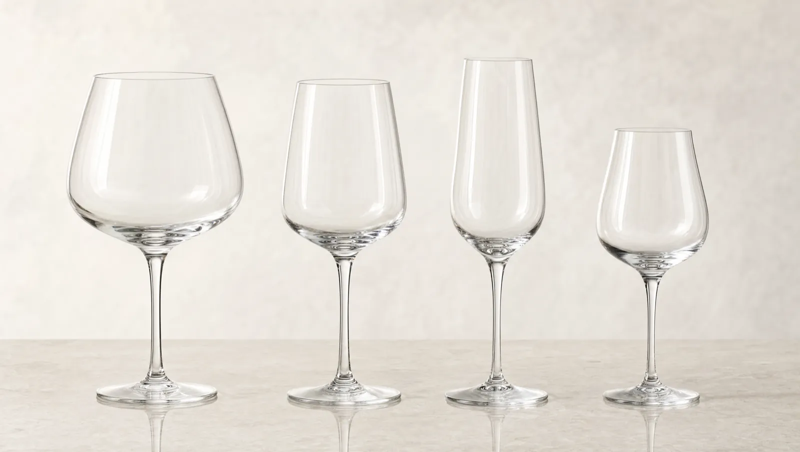Four empty crystal glasses of different bowl shapes lined up on a pale stone surface